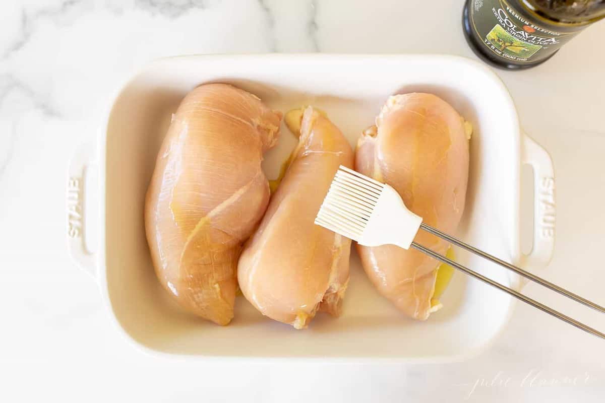 Three raw chicken breasts in a white baking dish, white basting brush on the top.