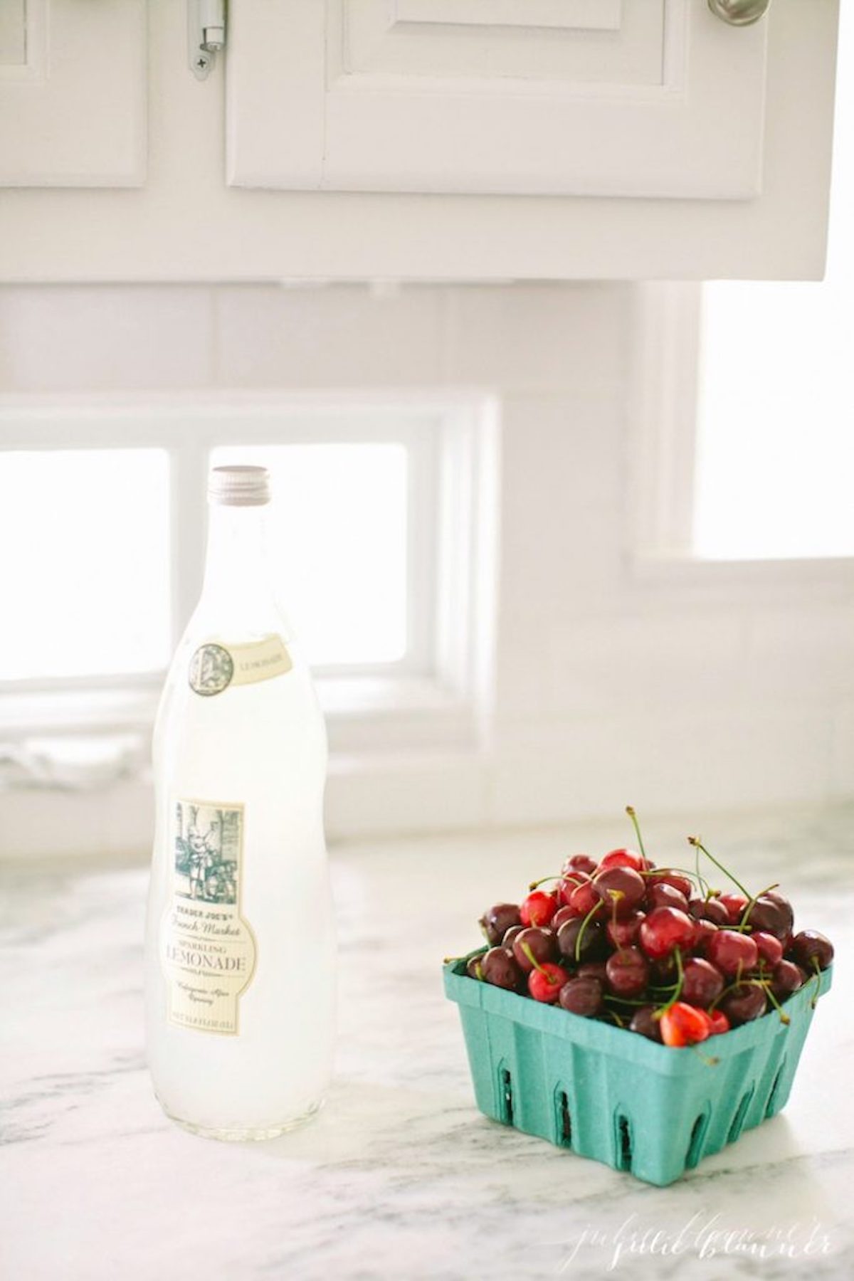 A white kitchen with mountain Danby marble countertops. A glass bottle of lemonade and a container of cherries are on the counters. 