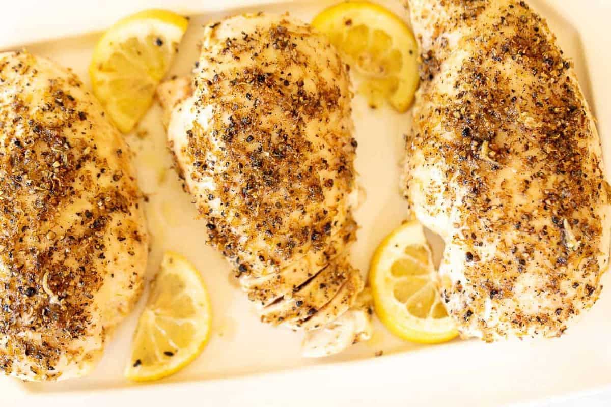 A white dish filled with lemon pepper chicken breasts, slices of lemon alongside.