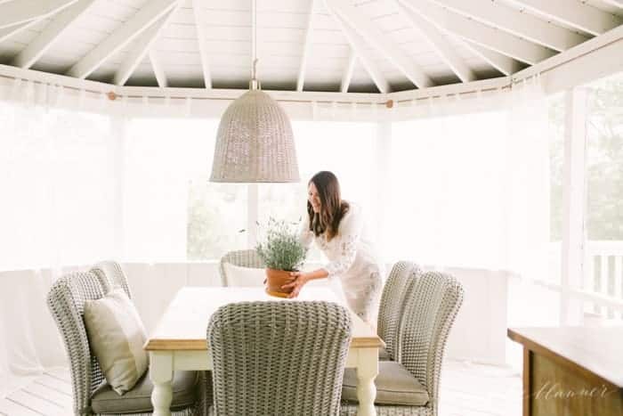 A table with woven chairs and light fixture above it in a screened in porch. Julie is putting a plant on the table. 