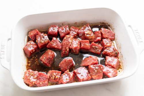 Raw marinated beef cubes, perfect for steak kabobs on the grill, arranged in a white baking dish, ready to be cooked.
