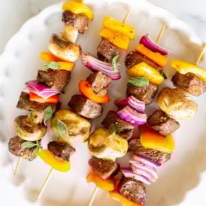 Three skewers with steak kabobs, grilled vegetables, mushrooms, and pieces of meat are arranged neatly on a white plate.