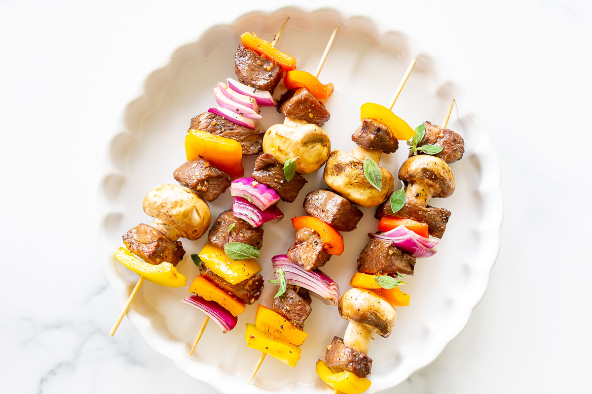 Three grilled skewers with marinated steak kabobs, mushrooms, red onions, and bell peppers served on a white plate.