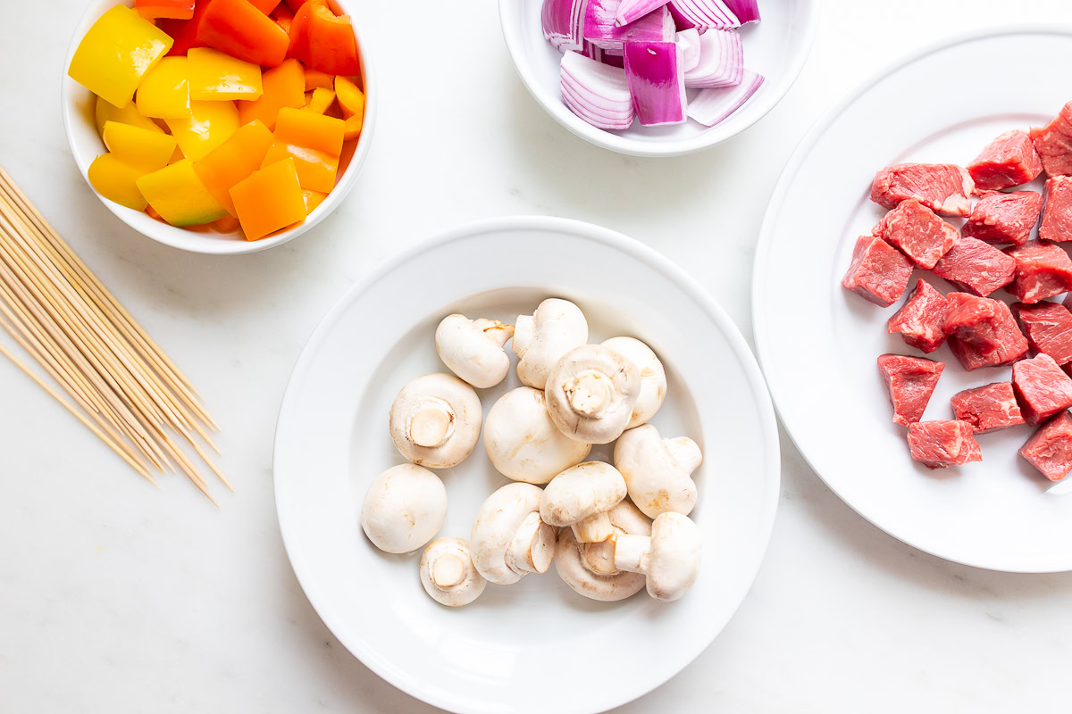 White bowls with yellow and orange bell pepper pieces, chopped red onions, white mushrooms, and diced steak for kabobs, along with wooden skewers on a white surface.