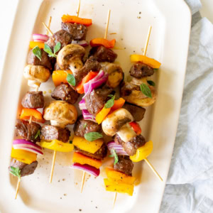Skewers with grilled steak kabobs, mushrooms, red onions, and red and yellow bell peppers arranged on a white rectangular plate.