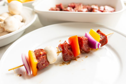 A skewer with marinated beef, bell peppers, red onion, and a mushroom on a white plate. In the background are bowls with raw mushrooms and steak kabob marinade.