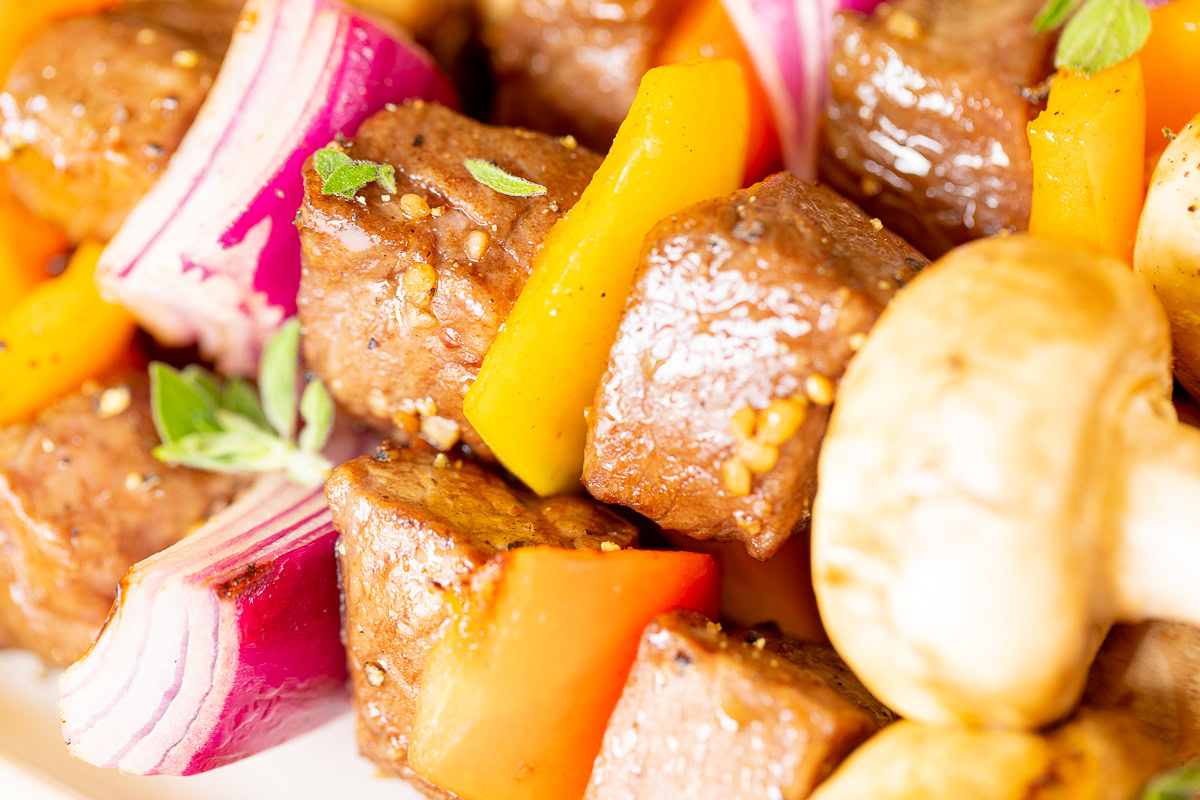 Close-up of skewered beef cubes, red onions, yellow and red bell peppers, mushrooms, and herbs on a plate. The vibrant assortment promises delightful flavors before they become succulent steak kabobs on the grill.