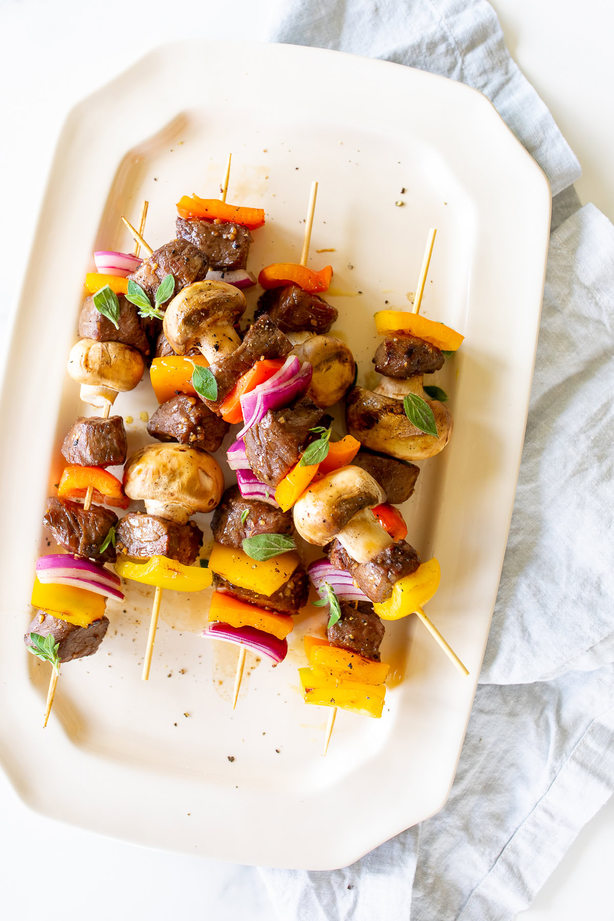 A white platter with skewers of steak kabobs, mushrooms, red onions, and bell peppers, garnished with fresh herbs, resting on a light blue cloth napkin.