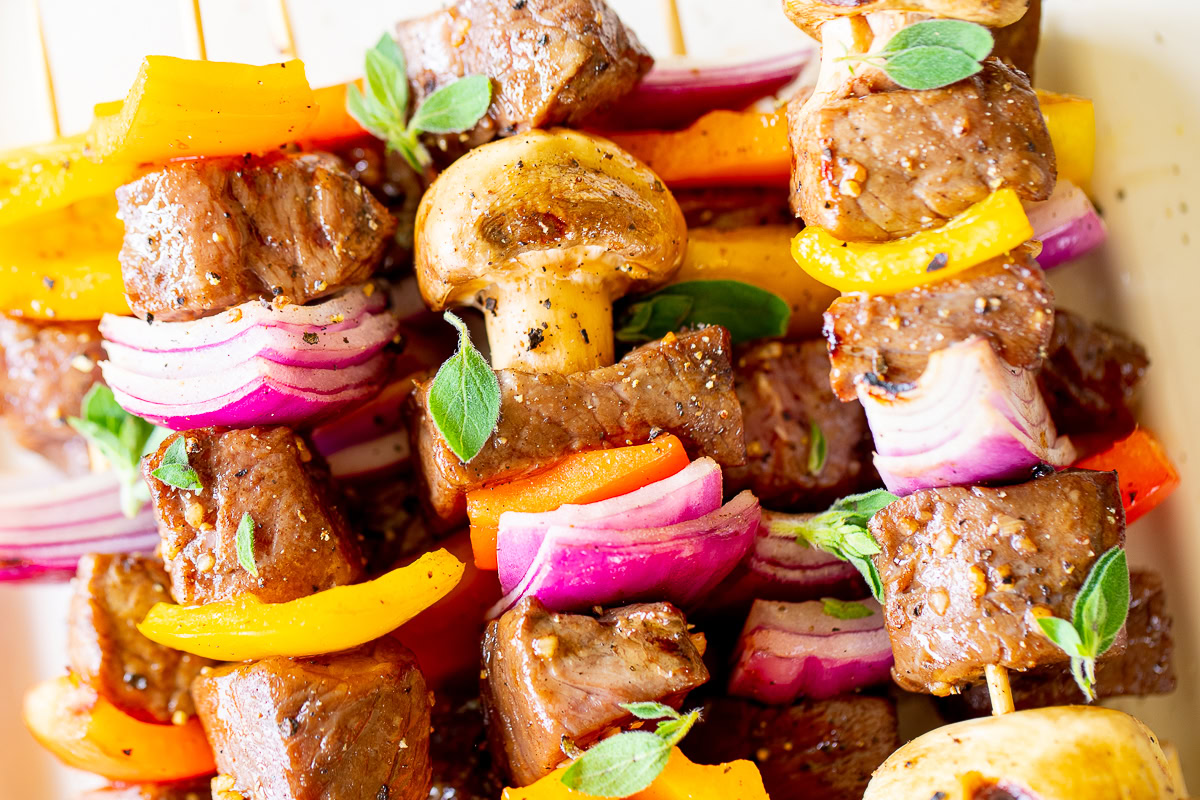 Close-up of colorful steak kabobs, featuring chunks of beef in a delicious steak kabob marinade, red onions, yellow and red bell peppers, and mushrooms, garnished with fresh herbs.