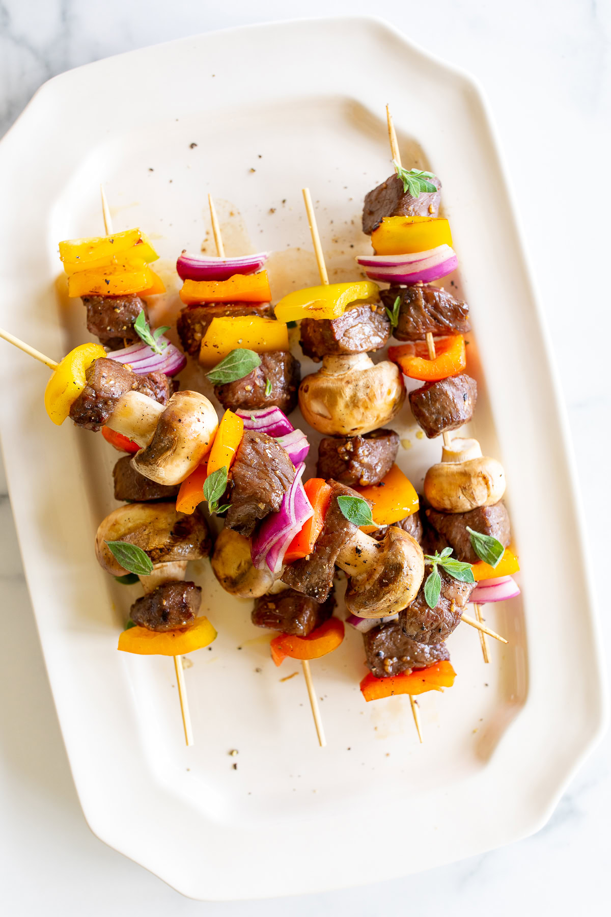 Grilled steak kabobs with beef, mushrooms, bell peppers, and red onions, garnished with herbs and served on a white rectangular plate.