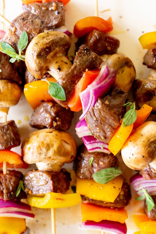 Close-up of colorful steak kabobs featuring chunks of beef, mushrooms, red and yellow bell peppers, and red onion, garnished with small green herbs.