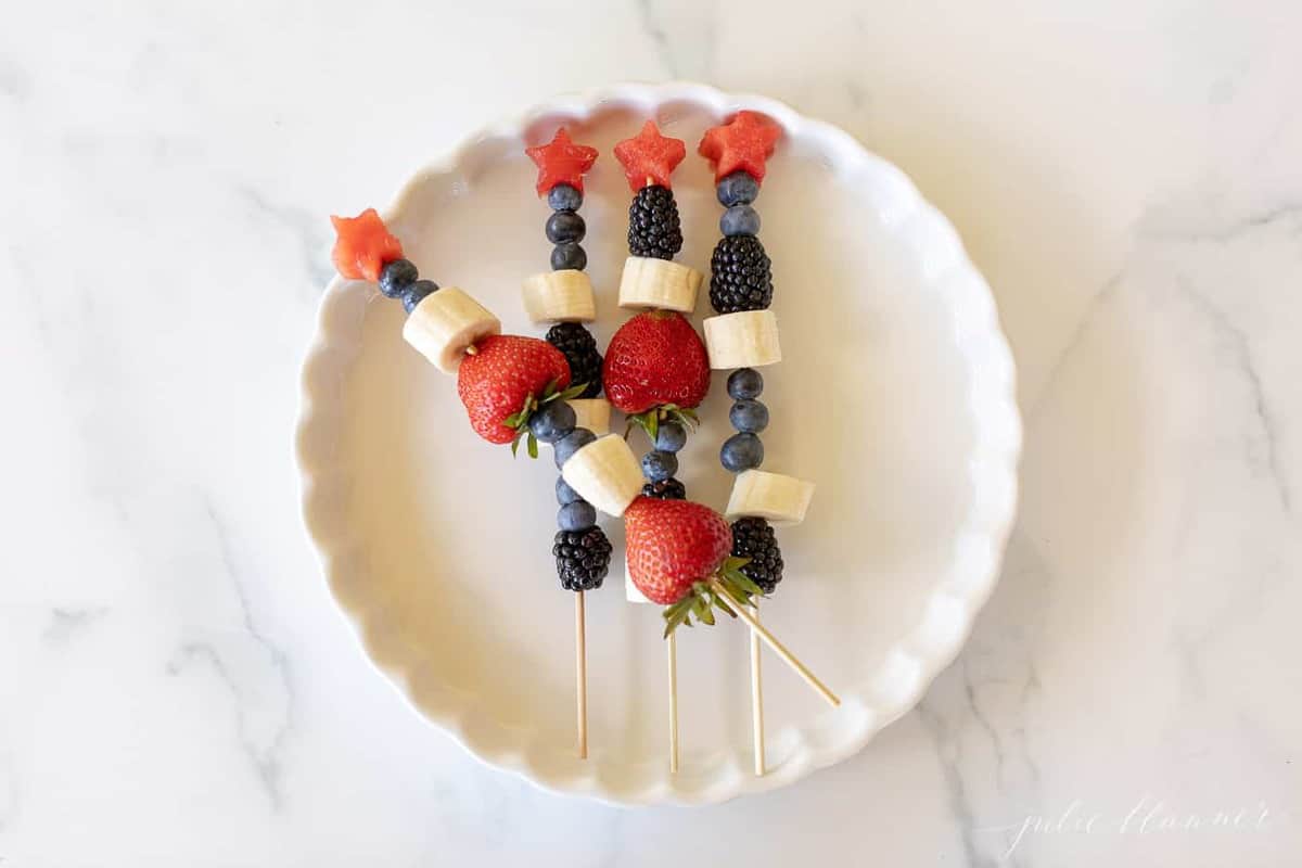 A round white platter on a marble surface, bamboo fruit skewers filled with berries on top.
