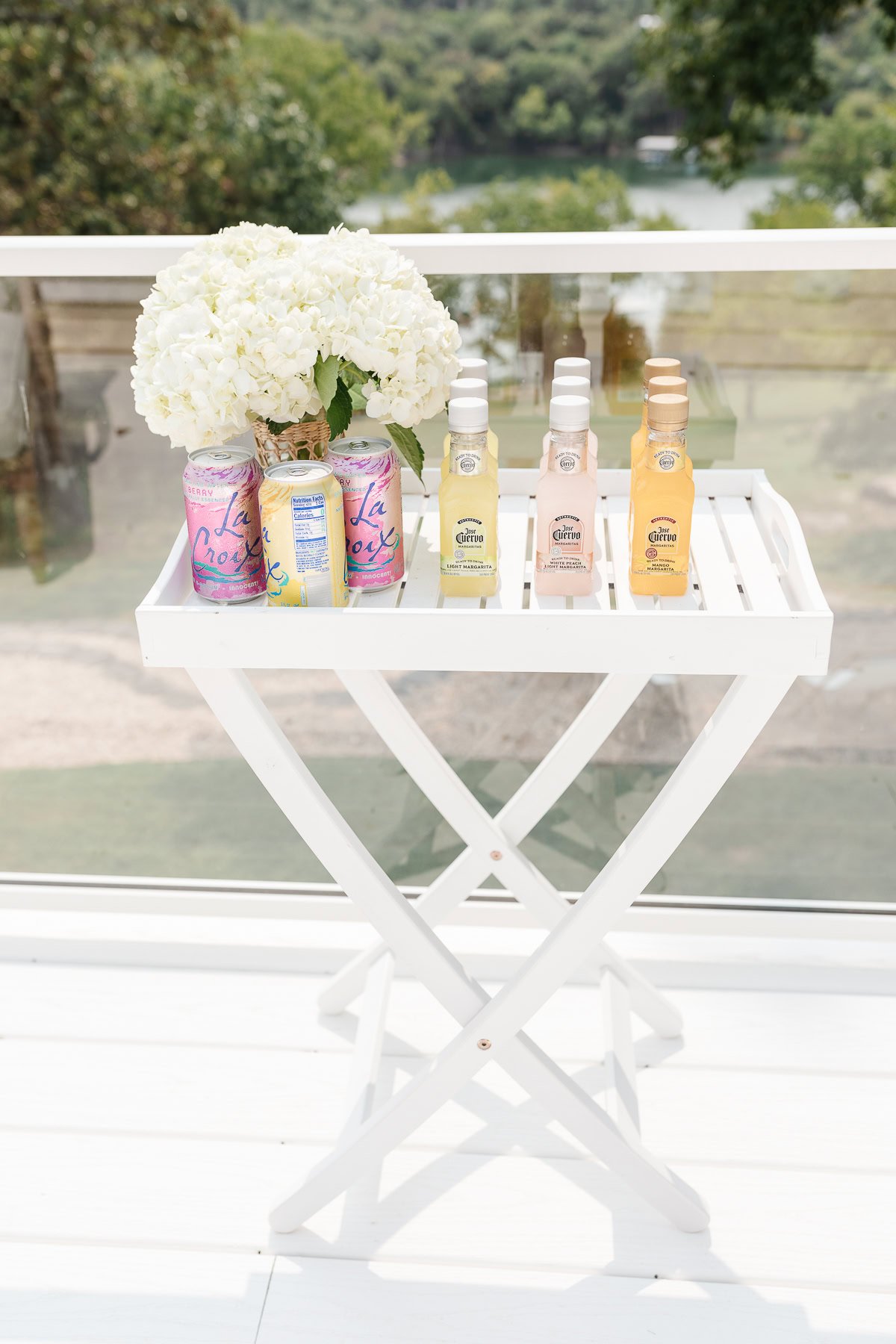 A white table with a bouquet of white flowers, three cans of LaCroix sparkling water, and three bottles of cocktail mixers sits on a porch overlooking a green landscape and body of water, all set on an elegant vinyl deck.