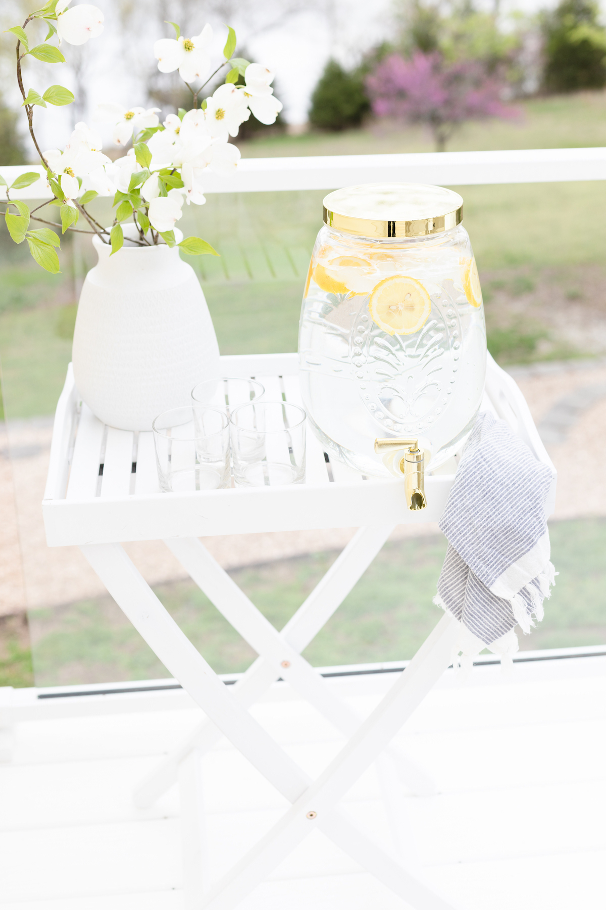 Lemon water dispenser, glasses, a white vase with flowers, and a striped towel on a white tray table outdoors on a stylish vinyl deck.