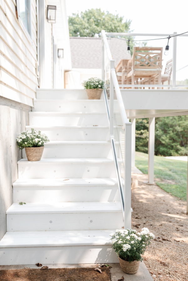 A white outdoor staircase leads to an elevated vinyl deck adorned with patio furniture. Flower pots with white blooming flowers are placed on the steps and at the base. Trees and a grassy area are visible.