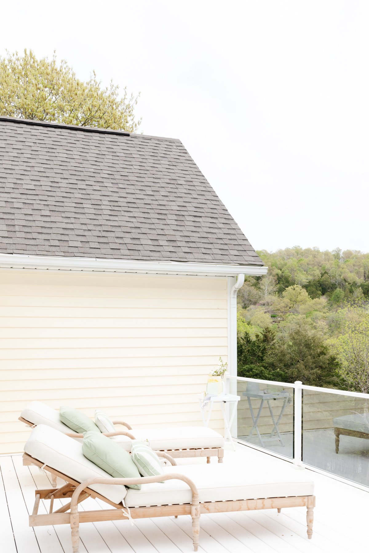 Two cushioned wooden lounge chairs with green pillows are on a white vinyl deck next to a house with light yellow siding. Trees are visible in the background.