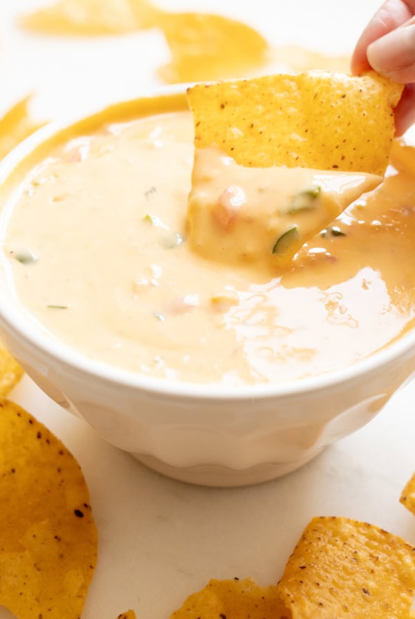 A hand dips a tortilla chip into a bowl of velveeta cheese dip on a white surface, surrounded by more chips.