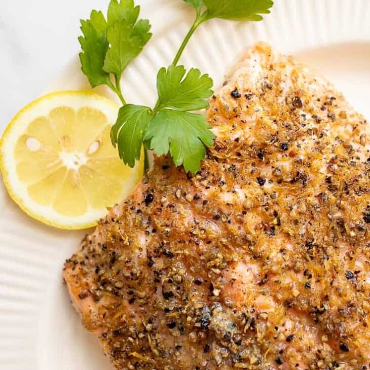 A white oval platter featuring a fillet of lemon pepper salmon, sliced lemons on the side.