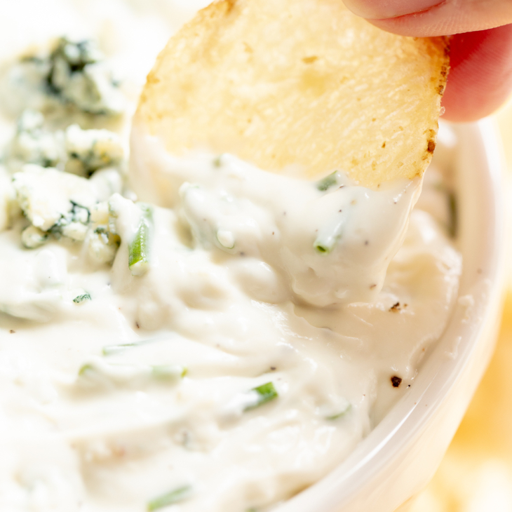 blue cheese dip in a white bowl, with fingers gripping a chip to dip into it.