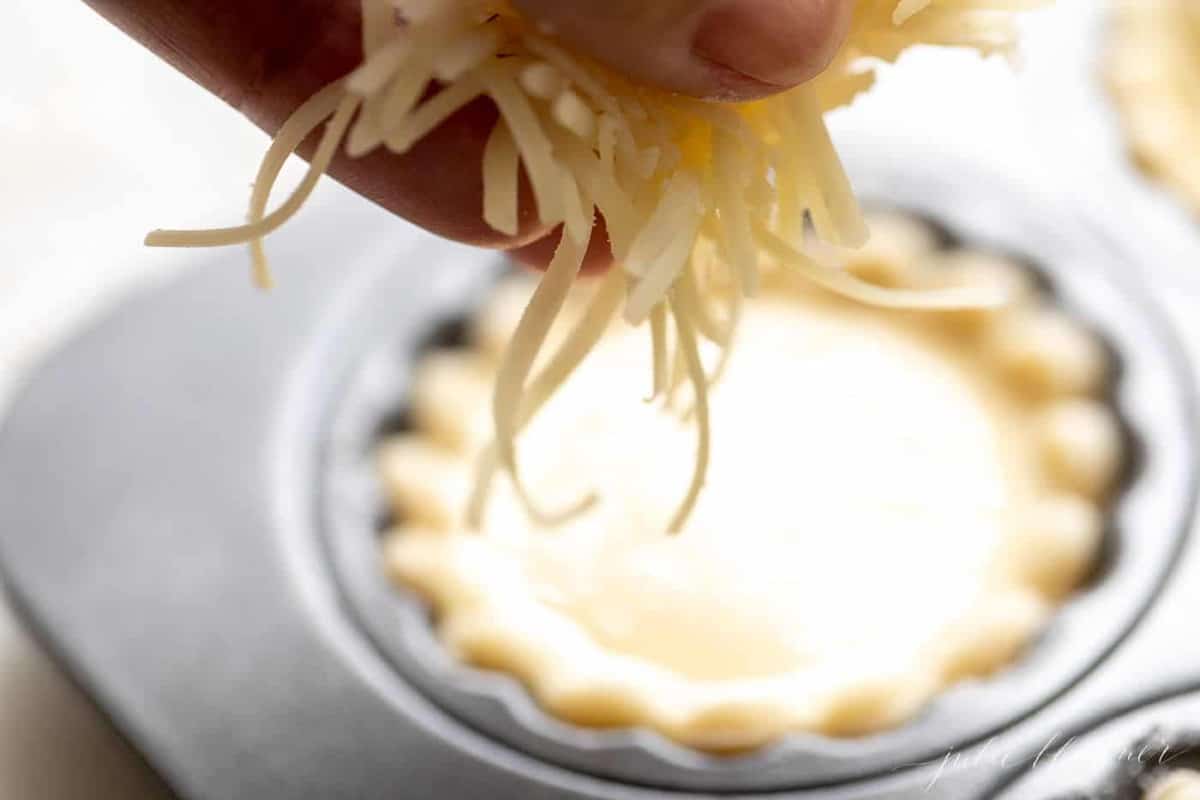 A hand preparing to place shredded cheddar cheese into a muffin cup