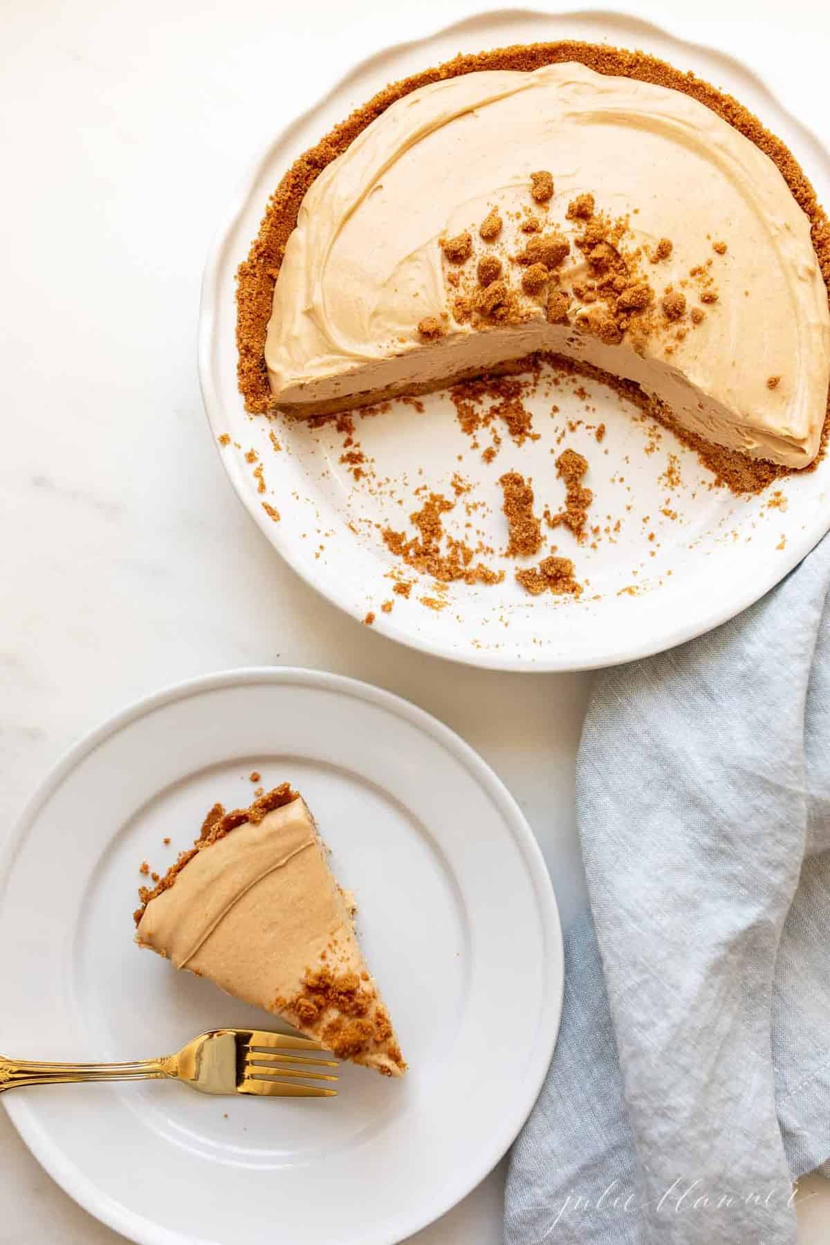 A cookie butter cheesecake on a white platter, with a plate to the side with a slice of cheesecake and fork.