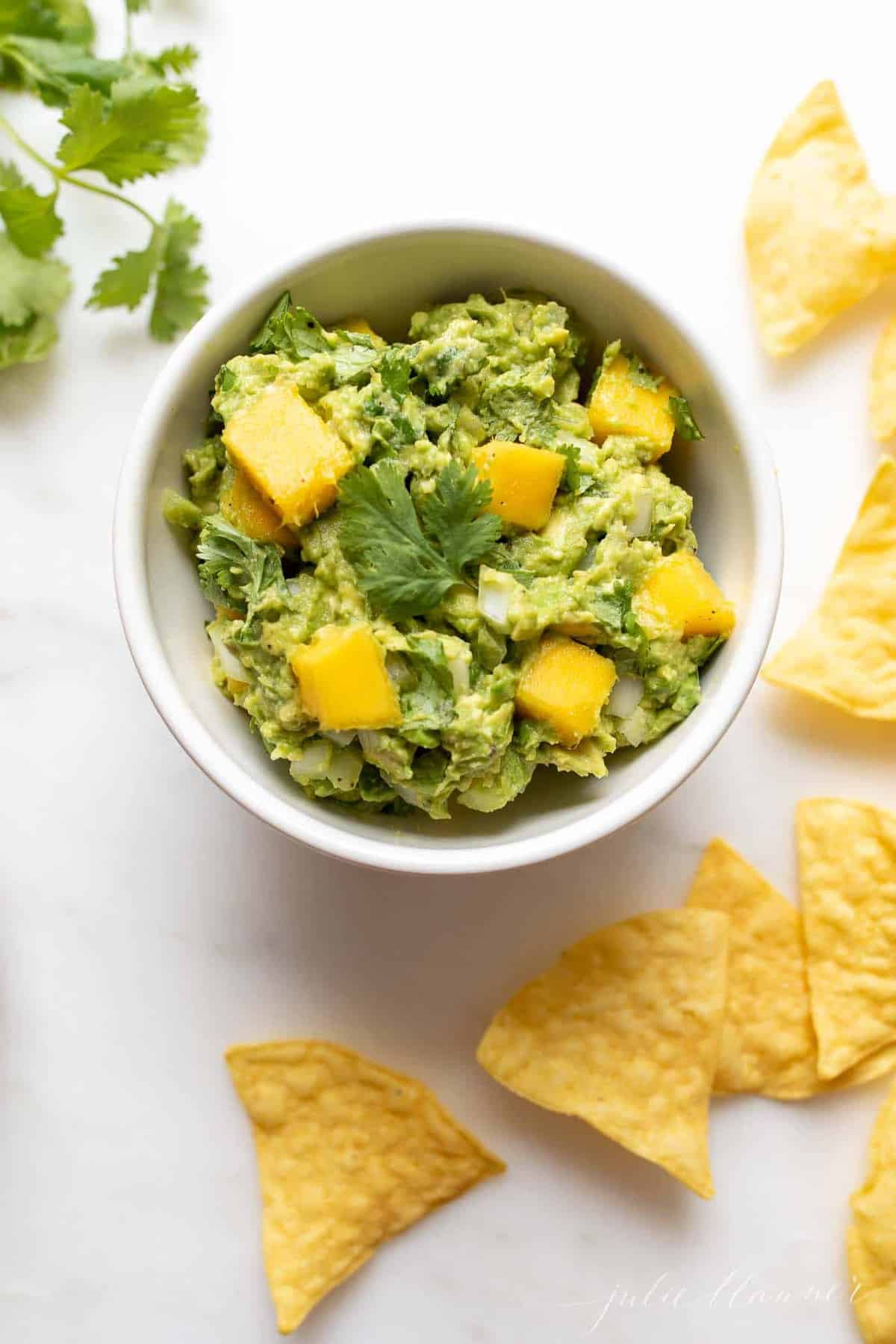 A white bowl on a marble surface full of mango guacamole, chips to the side.