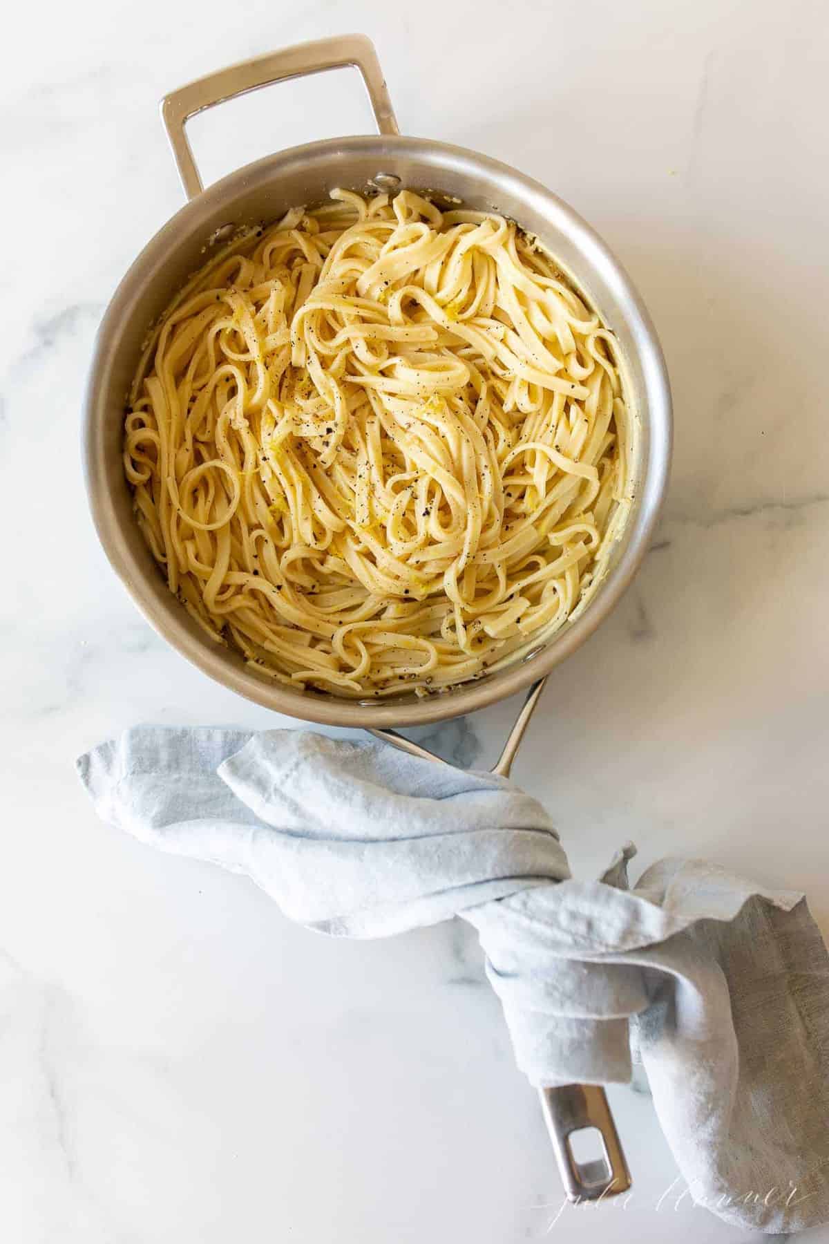 A silver pan filled with cooked fettuccine, blue linen towel wrapped around handle