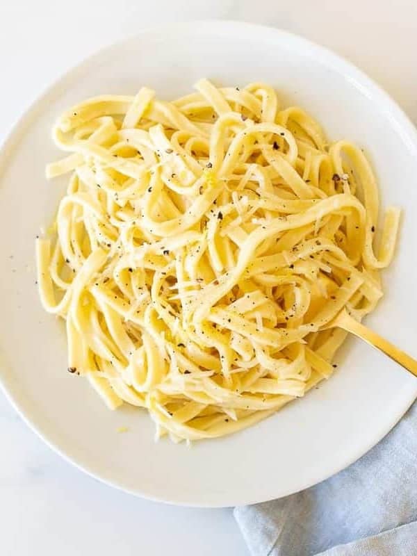 A white plate with a bowl full of lemon pepper pasta, gold fork to the side.