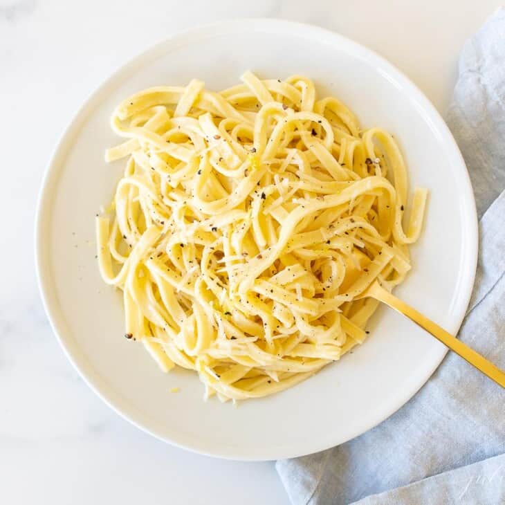 A white plate with a bowl full of lemon pepper pasta, gold fork to the side.