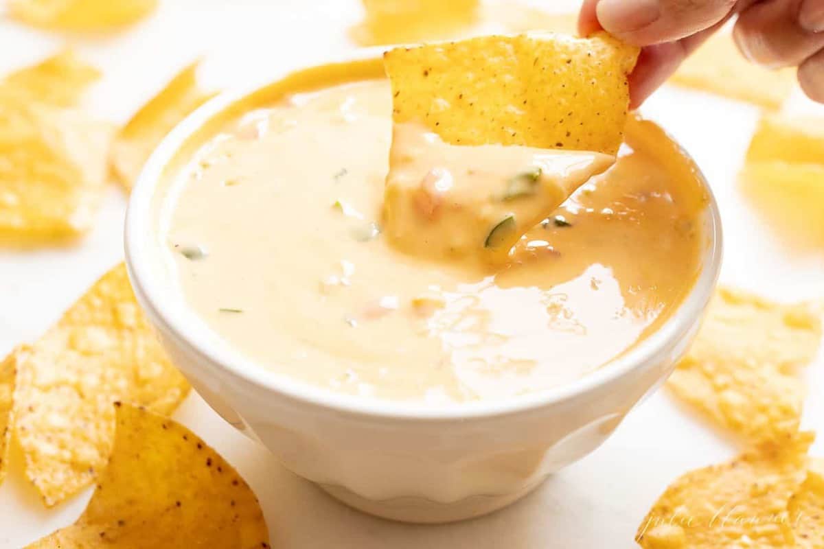 A hand reaching into a bowl full of Velveeta cheese dip, chips surrounding the bowl.