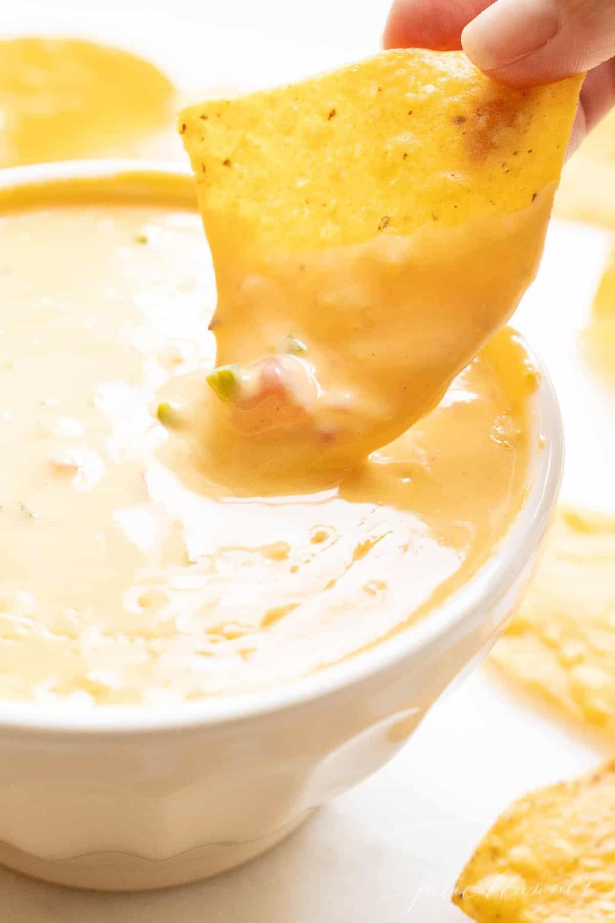 A hand reaching into a bowl full of Velveeta cheese dip, chips surrounding the bowl.