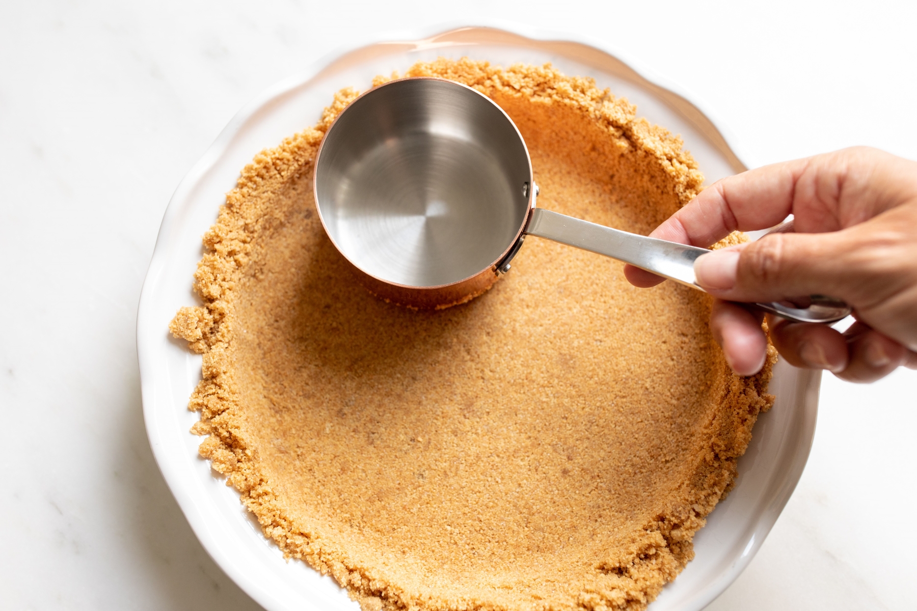 A hand with a copper measuring cup on a homemade graham cracker crust.