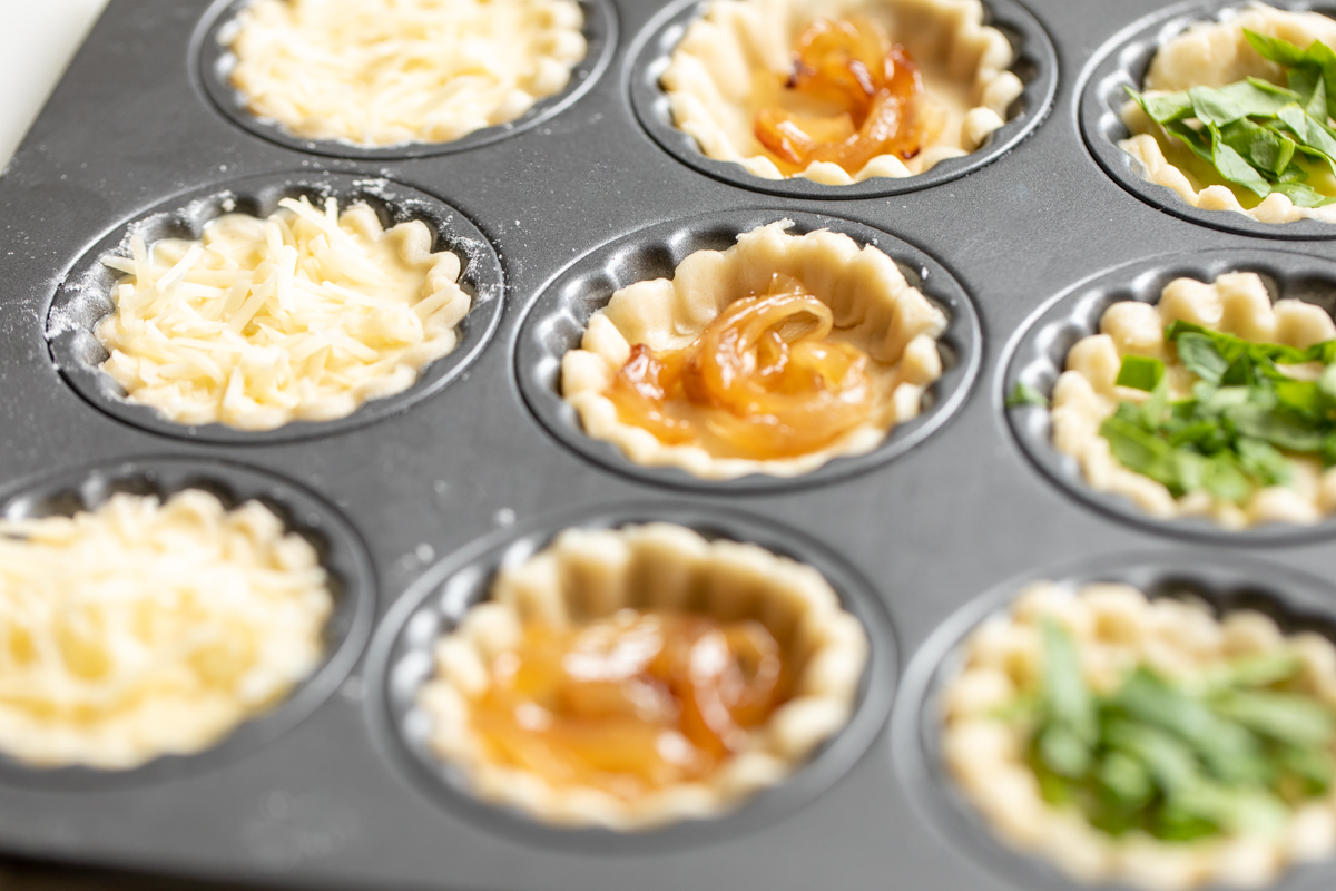 A variety of mini cheese tarts in a muffin tin, prior to baking.