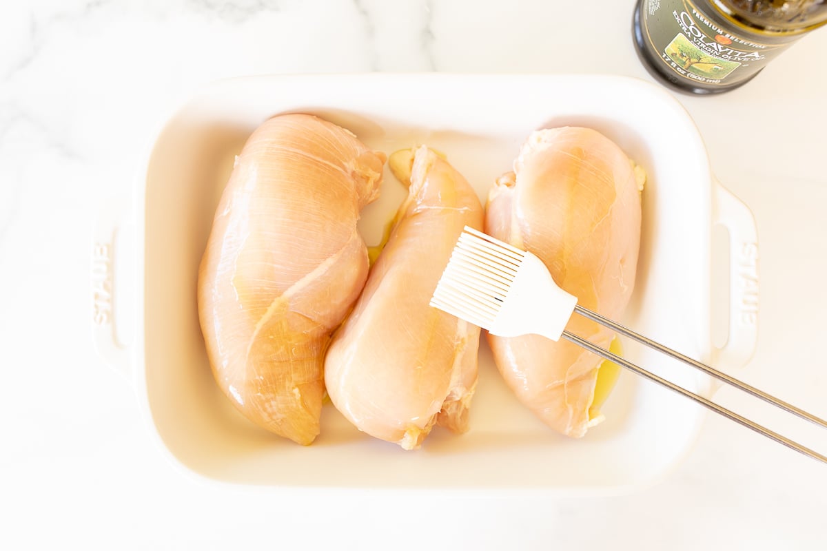 Raw chicken breasts in a white baking dish, with a brush adding olive oil on top.