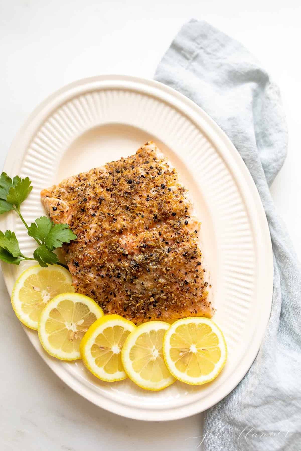 A white oval platter featuring a fillet of lemon pepper salmon, sliced lemons on the side.