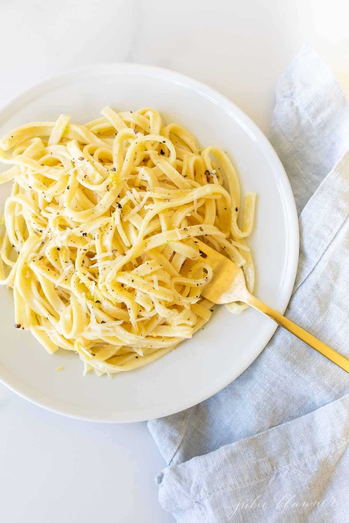 A white plate with a bowl full of lemon pepper pasta, gold fork to the side.