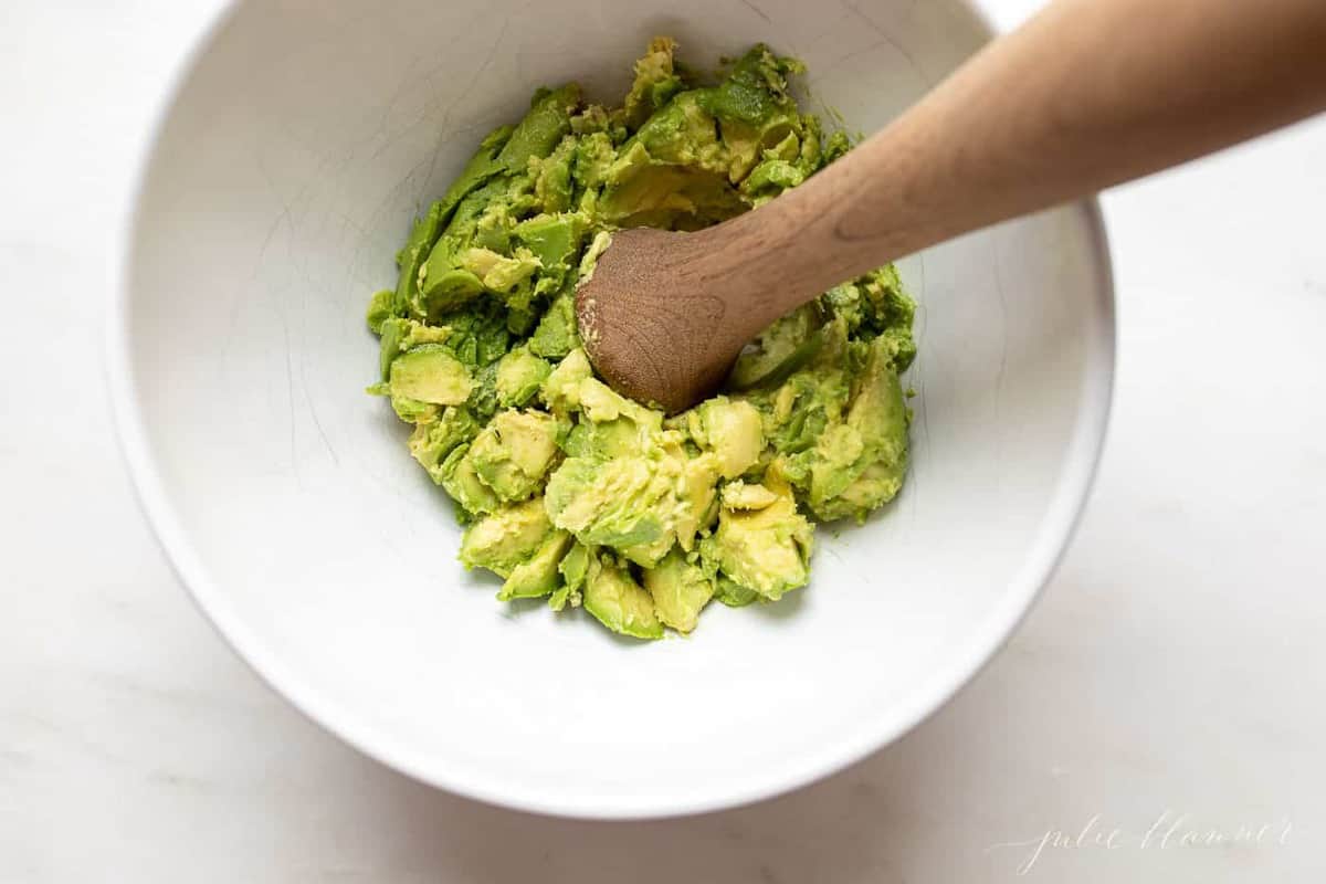 Looking into a white bowl full of avocado chunks, wooden pestle to the side.