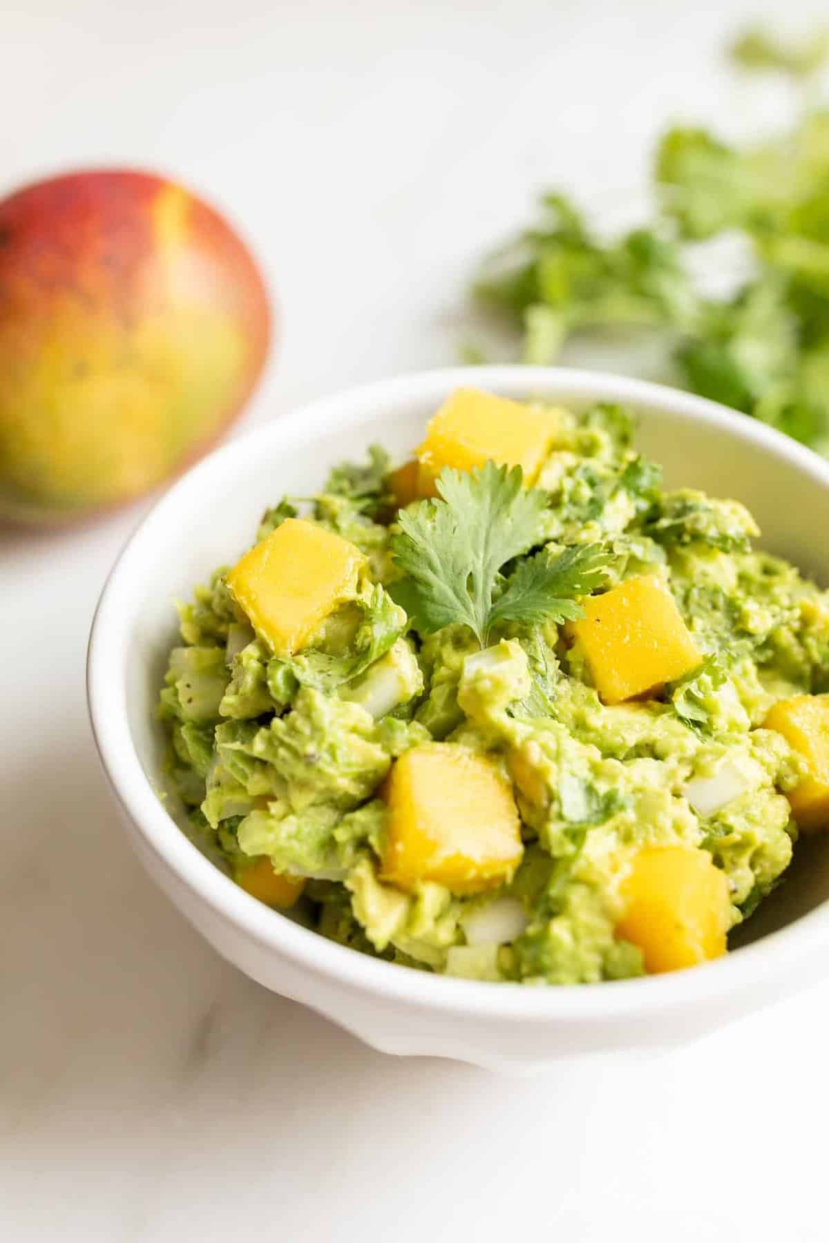 A white bowl on a marble surface full of mango guacamole, chips to the side.