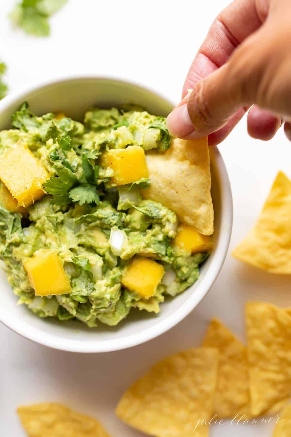 A white bowl on a marble surface full of mango guacamole, chips to the side and a hand reaching into the dip bowl.