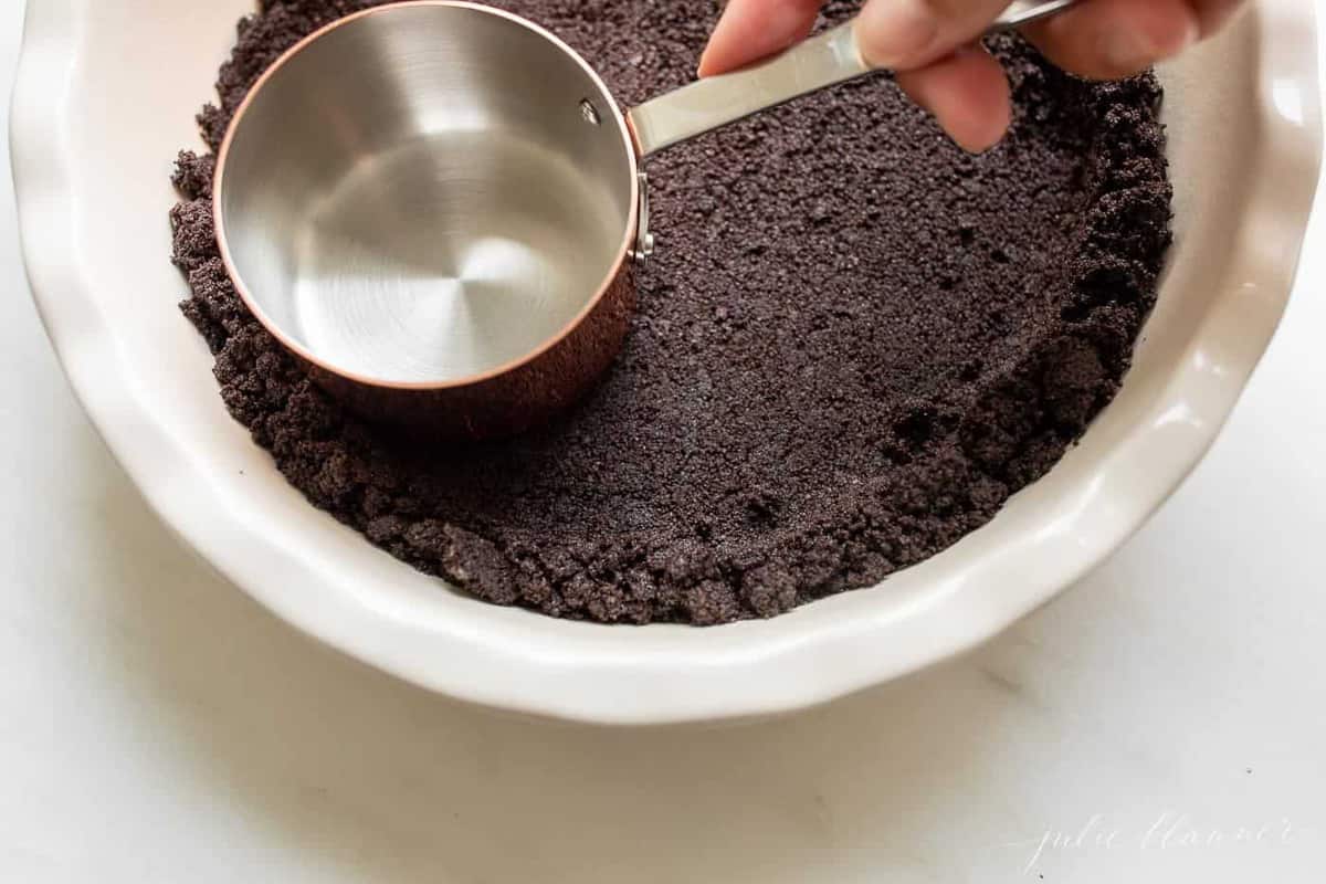 A hand pressing oreo cookie crumbs into a pie dish with a measuring cup for an oreo cookie crust.
