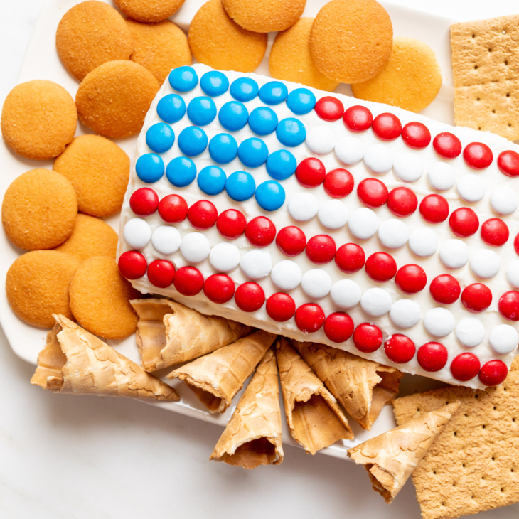 A red white and blue snack made of Funfetti dip in a flag shape, on a plate of cookies and crackers.