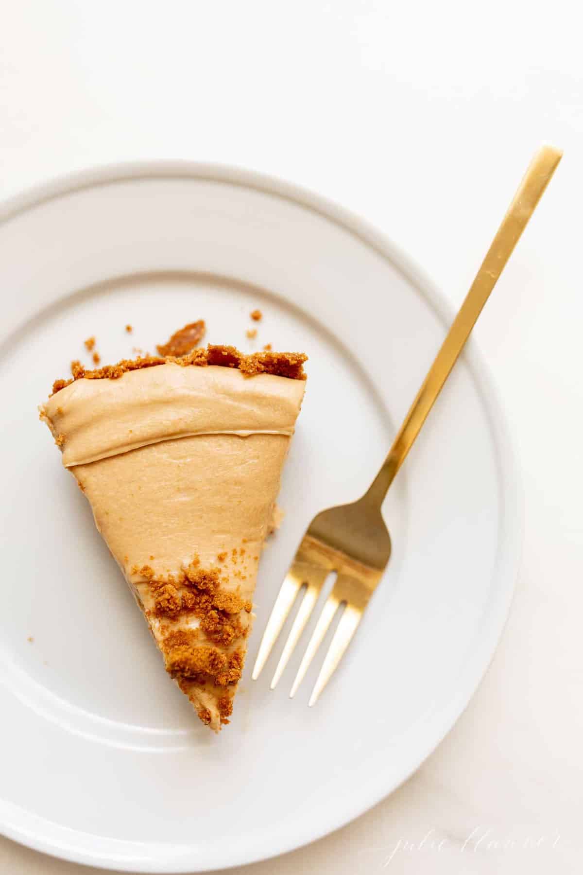 A white surface with a white plate with a slice of cookie butter cheesecake, gold fork to the side.