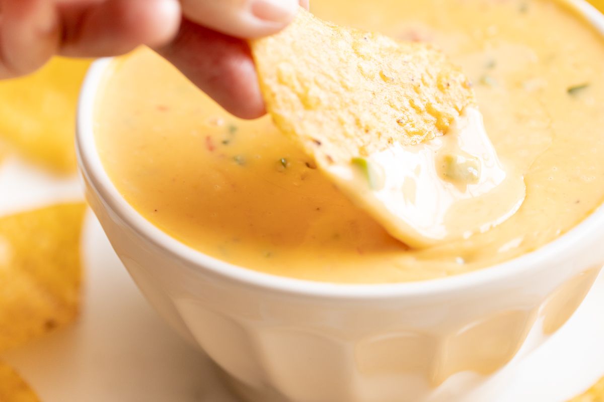 A hand dipping a tortilla chip into a white bowl of Velveeta cheese dip.