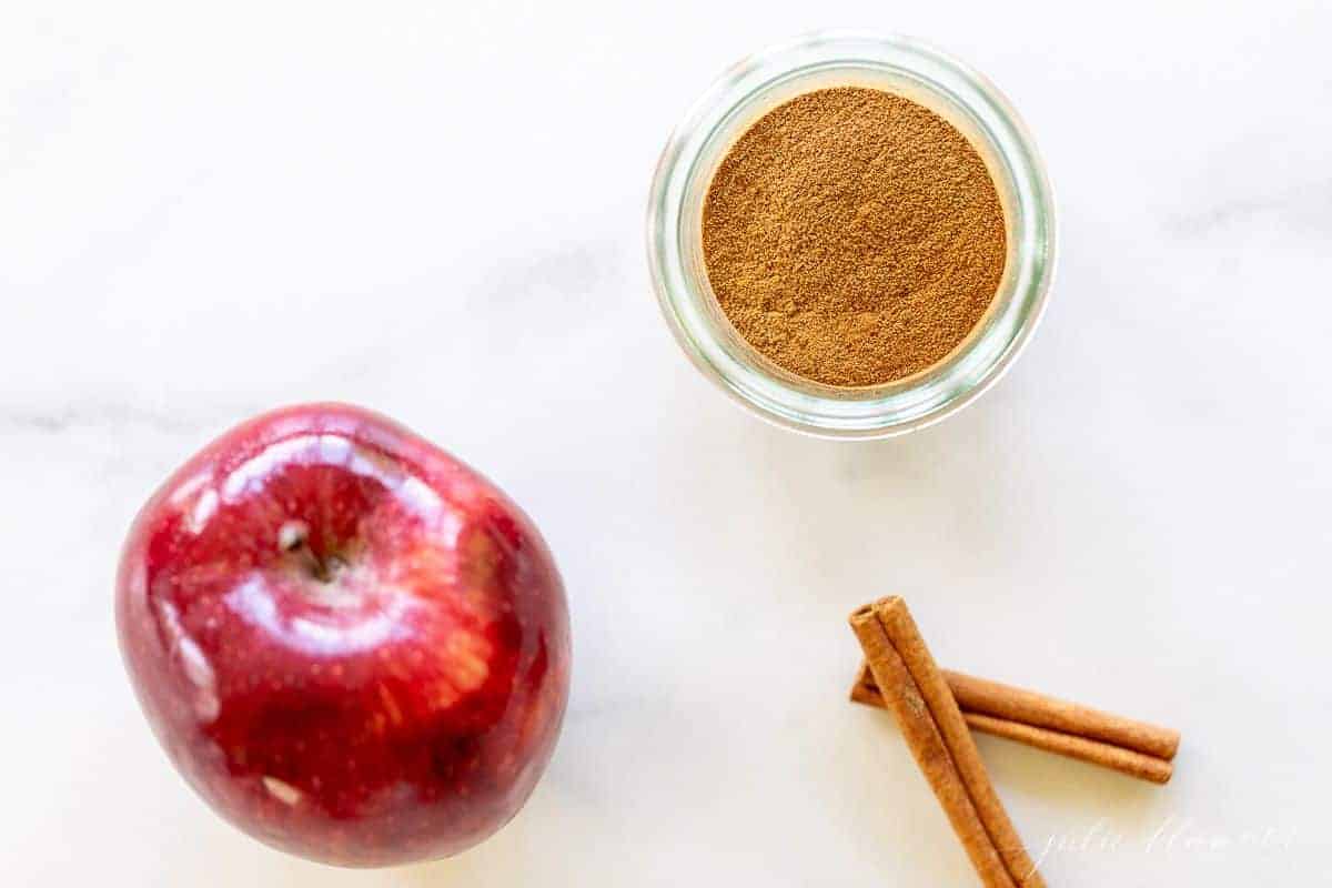 A marble surface with a small jar of apple pie spice, cinnamon sticks and an apple to the side.
