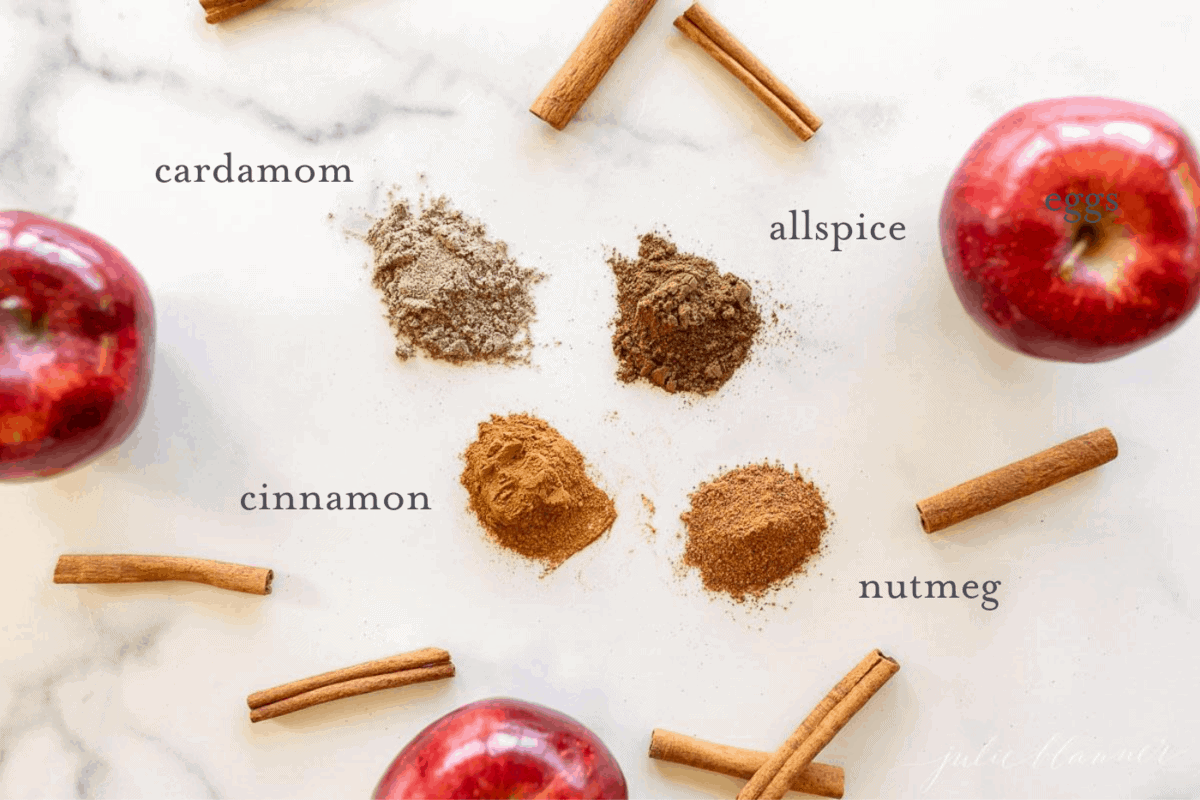 A marble surface filled with seasoning piles, cinnamon sticks and apples to make an apple pie spice recipe.
