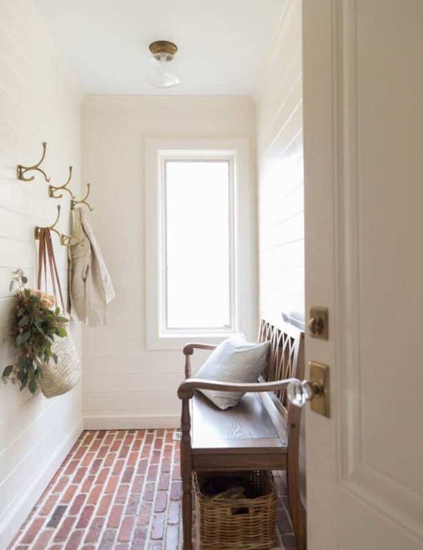 A white shiplap mudroom with thin brick flooring, brass hooks on the walls and a wooden bench.