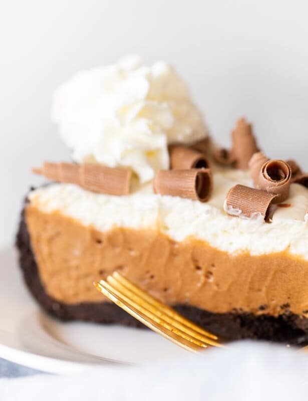 A slice of french silk pie topped with whipped cream and chocolate shavings on a white plate, gold fork to the side.