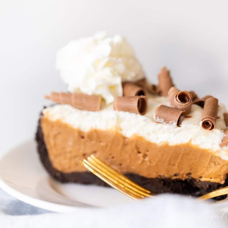 A slice of french silk pie topped with whipped cream and chocolate shavings on a white plate, gold fork to the side.