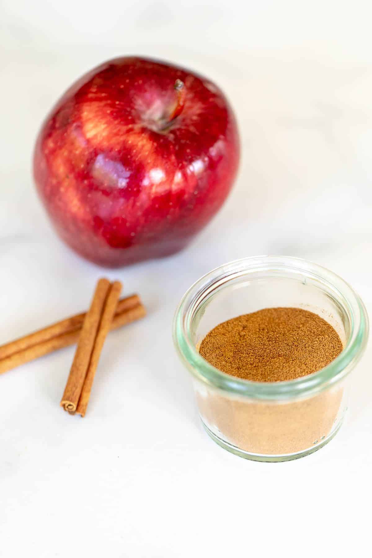 A marble surface with a small jar of apple pie spice, cinnamon sticks and an apple to the side.