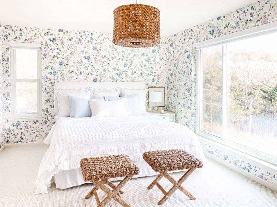 A bedroom with patterned floral slightly dated wallpaper, updated with fresh white bedding and pretty textures with a large window.